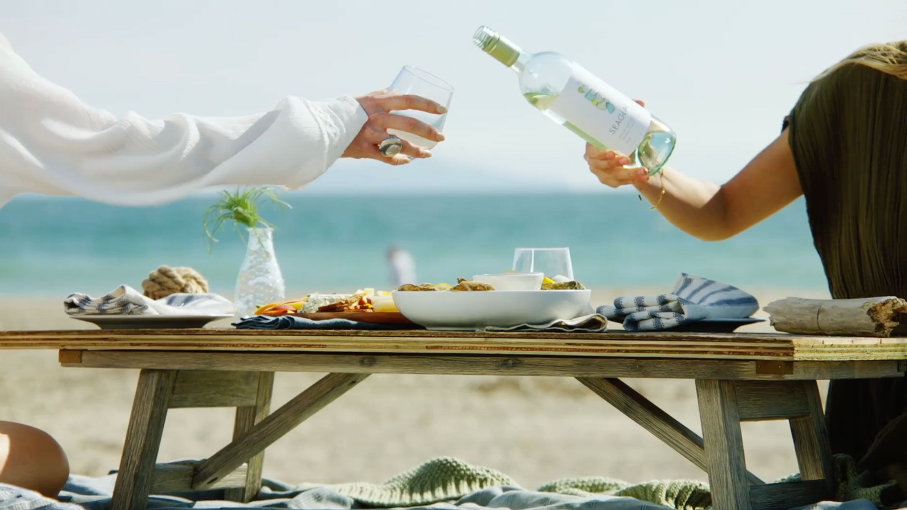 One person holds a wine glass, while another leans across a low wooden table of food to pour a bottle of white wine into it. The table holds picnic food and the ocean is in the background