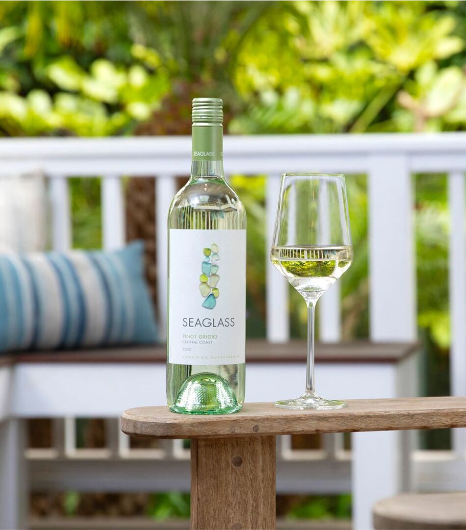 A bottle and glass of Seaglass of Pinot Grigio on the wooden arm of a chair with a white fence, wooden bench, and garden in the background