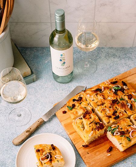 Sliced focaccia lies on a wooden cutting board, next to a bread knife, two glasses of white wine, and a bottle of Seaglass Central Coast Sauvignon Blanc